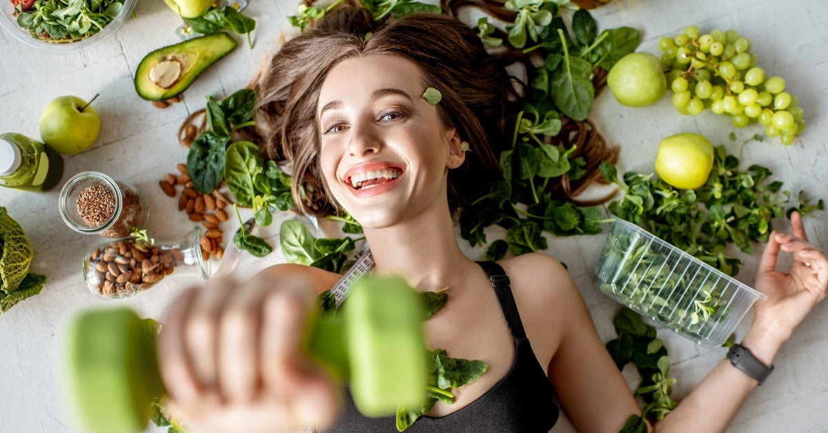 Femme sportive souriante tenant des haltères verts, entourée d’aliments sains et légumes frais