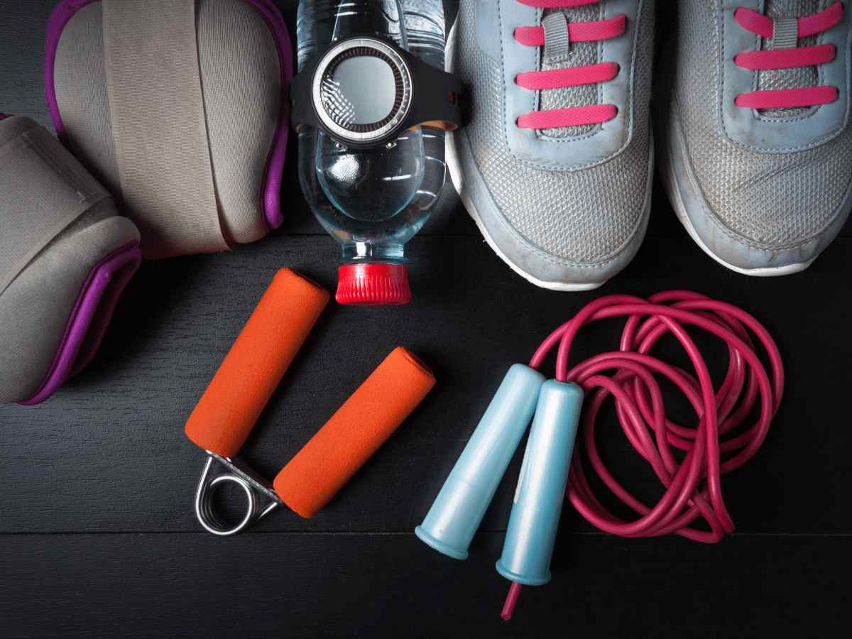 Accessoires de sport essentiels – chaussures, gourde, corde à sauter et haltères posés sur un sol de gym.